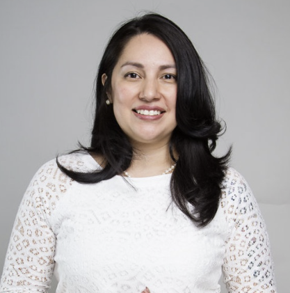 A medium brown skin tone Chicana woman, with shoulder length dark brown hair, is smiling at the camera. She is wearing a white lace blouse and is standing against a gray background.